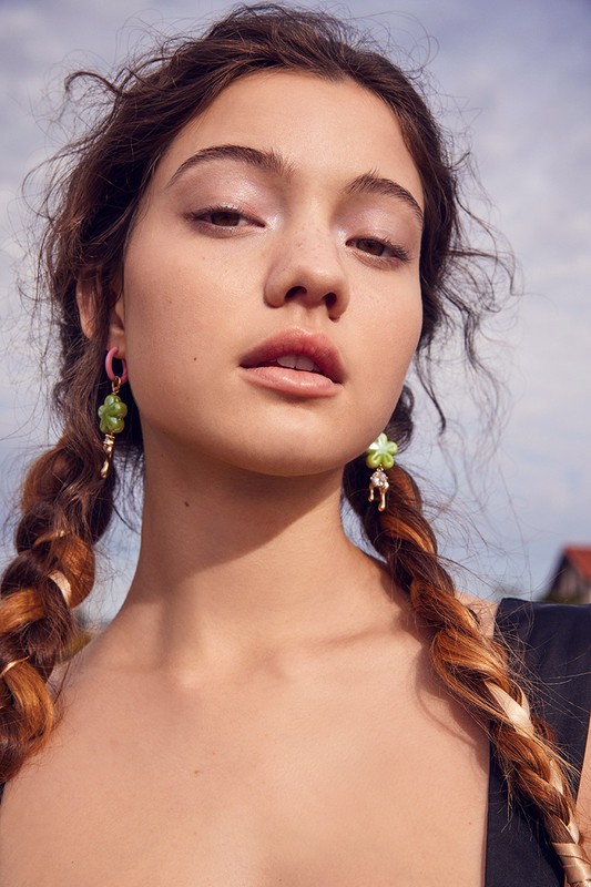 Close-up of a woman with pigtails, wearing green floral earrings, posing outdoors against a sky background, beauty and fashion.