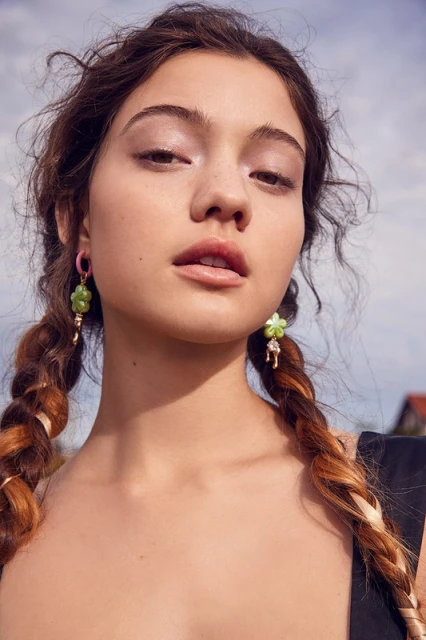 Close-up of a woman with pigtails, wearing green floral earrings, posing outdoors against a sky background, beauty and fashion.