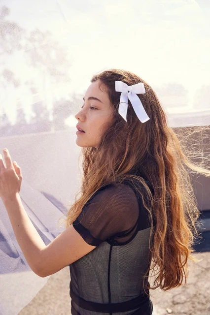 Portrait of a young woman with long wavy hair wearing a sheer top and a white bow in her hair, standing outdoors.