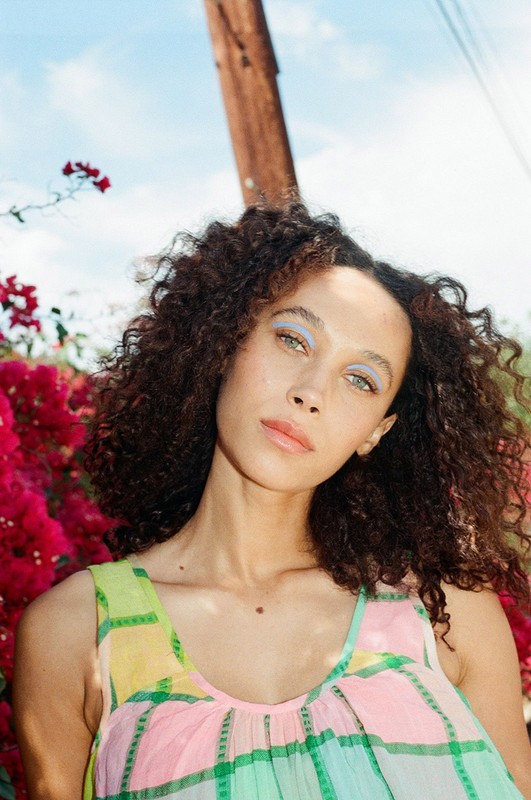 Portrait of a beautiful African-American woman with curly hair, wearing a multicolored dress, posing in front of bougainvillea and a blue sky.