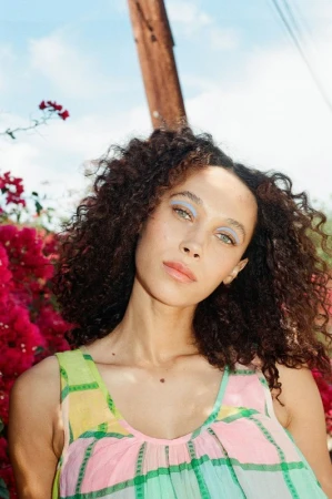 Portrait of a beautiful African-American woman with curly hair, wearing a multicolored dress, posing in front of bougainvillea and a blue sky.