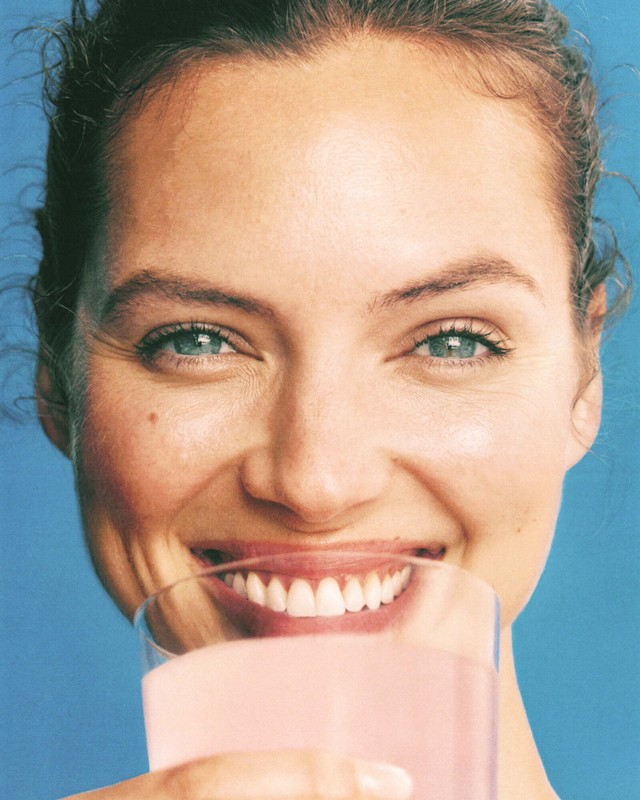Close-up of a smiling woman with green eyes holding a pink liquid in a clear glass against a light blue background, beauty shot.