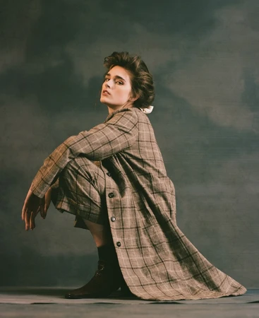 Model in a plaid trench coat crouches against a mottled dark studio backdrop with styled voluminous hair.