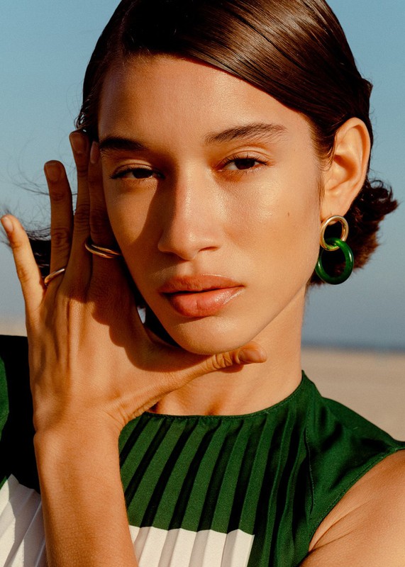 Close-up shot of a young adult female model with dark hair, wearing earrings, and a dark green and white striped top against a blue sky.
