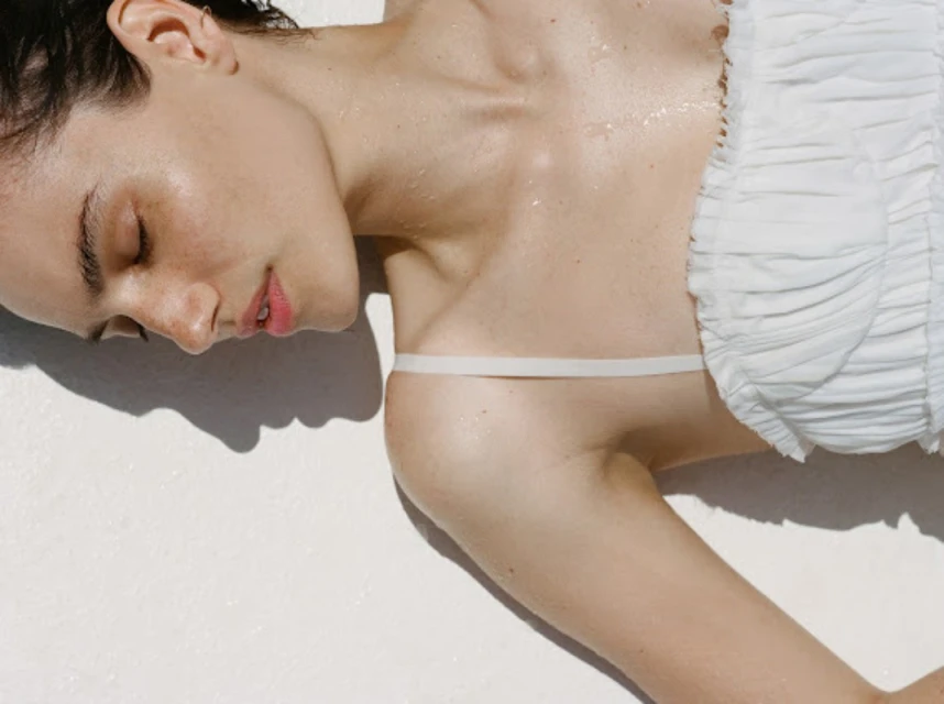 Close-up of an adult woman with short hair lying on a white surface, wearing a white ruffled top and a single white strap.