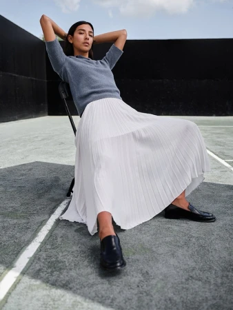 A woman in a blue short-sleeve top and white pleated skirt seated on a chair outdoors on a court with black walls.