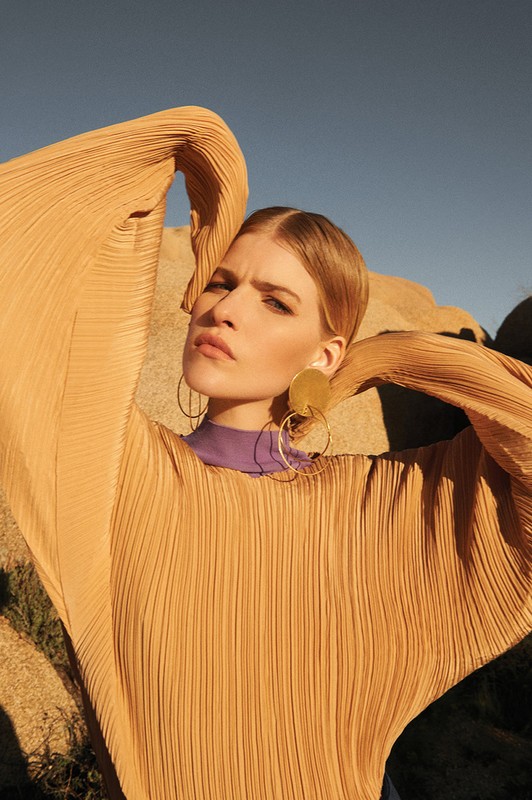 Woman in draped tan top, gold earrings poses outdoors against rock formations under blue sky in fashion shot, portrait orientation.