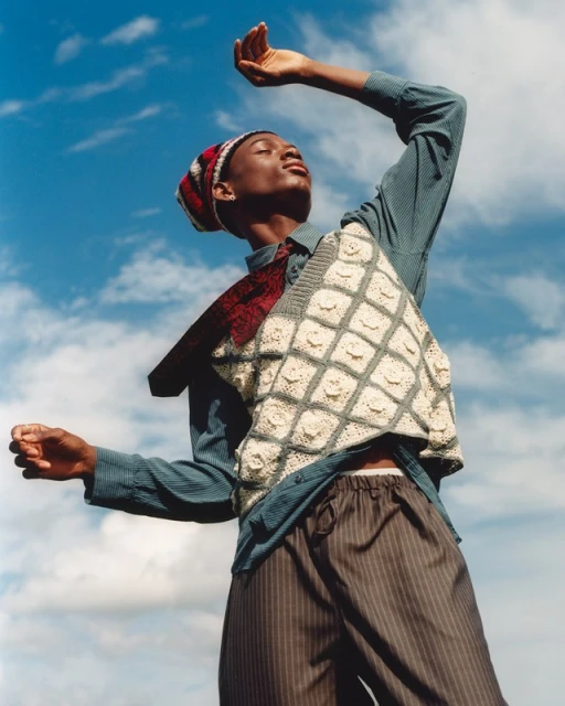 Young adult with upraised arms in a knitted vest, hat, and pinstripe pants set against a blue sky backdrop on a sunny day.