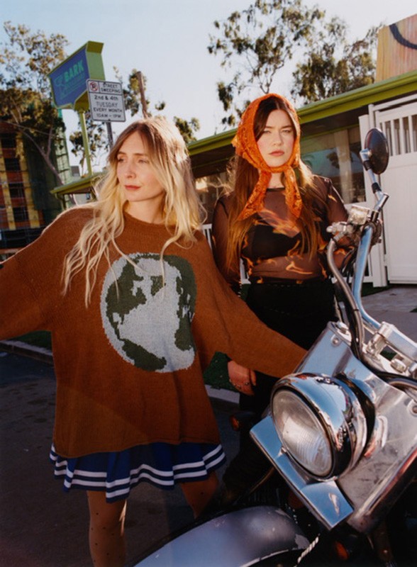 Two young women pose outdoors next to a vintage motorcycle on a sunny day; one wears a world-motif sweater, the other a headscarf.