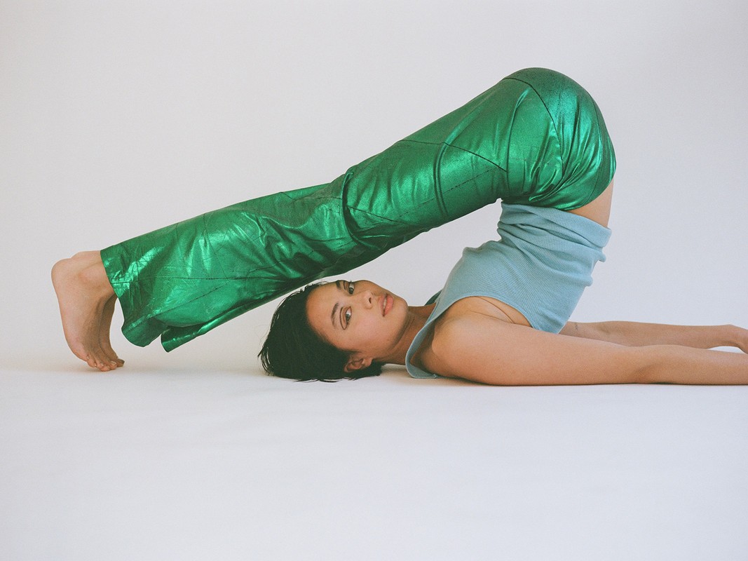 Woman wearing green metallic pants lies on her back, legs raised in the air in a shoulder stand position against a plain white backdrop.