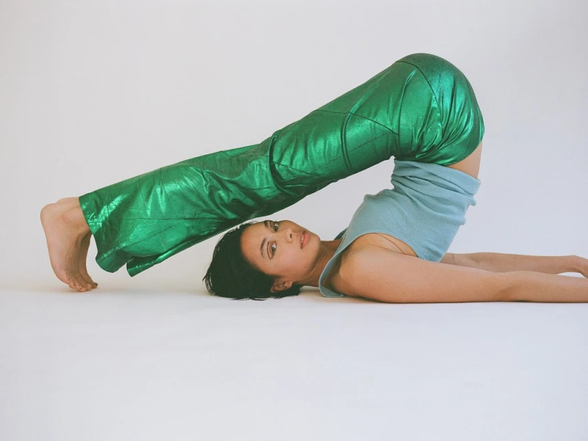 Woman wearing green metallic pants lies on her back, legs raised in the air in a shoulder stand position against a plain white backdrop.