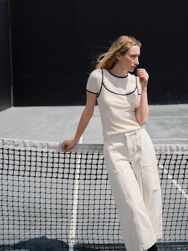 A slender woman stands at a tennis net, wearing an ivory tee with dark trim and white pants, against a solid dark background.