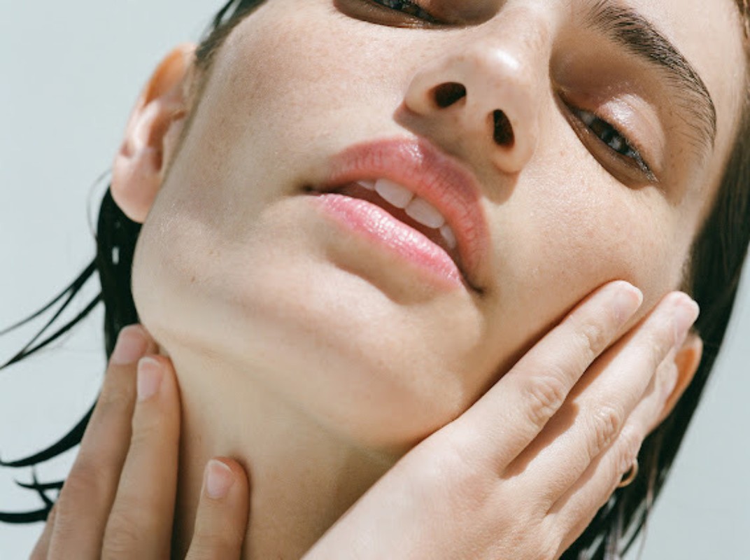 Close-up beauty portrait of a woman with wet hair touching her jawline and neck, featuring natural makeup.
