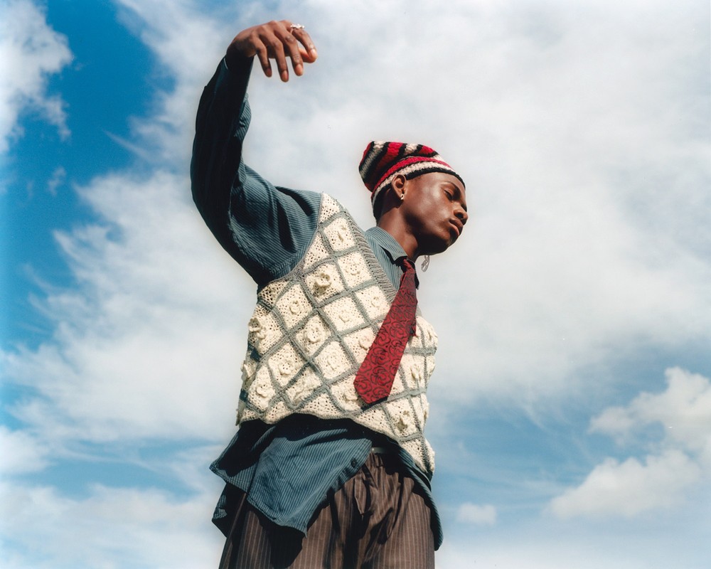 Fashionable young adult poses against a blue sky wearing a knit hat, vest, striped shirt, and tie in an outdoor setting.