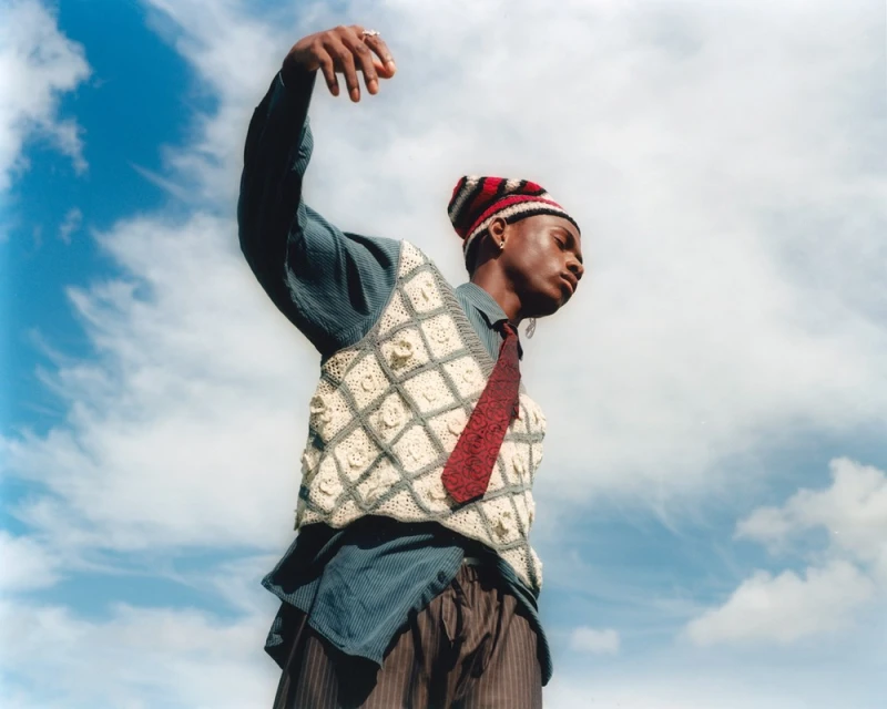Fashionable young adult poses against a blue sky wearing a knit hat, vest, striped shirt, and tie in an outdoor setting.