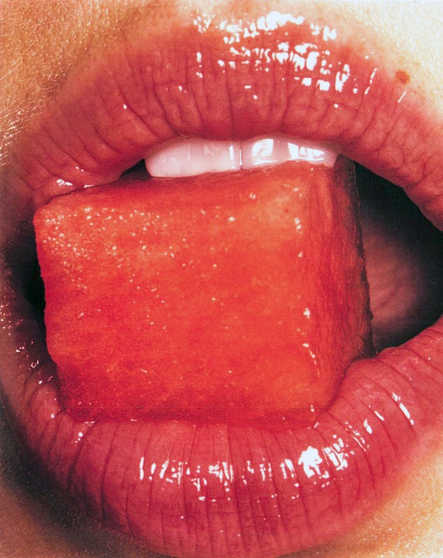Close-up of a person's mouth with red lipstick, holding a red watermelon cube on their tongue; teeth are visible.