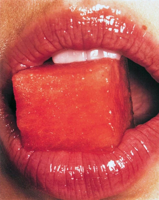 Close-up of a person's mouth with red lipstick, holding a red watermelon cube on their tongue; teeth are visible.