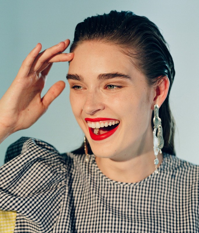 Portrait of a smiling woman with wet-look hair, red lipstick, and distinctive earrings wearing a black and white checkered top.