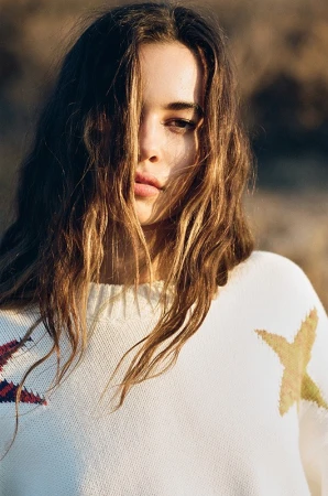 Portrait of a woman with long tousled brown hair wearing a white knit sweater with a star pattern under natural sunlight.