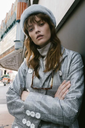 Woman in a grey plaid coat and beret stands on a city street with an old theater in the background, wearing glasses. 