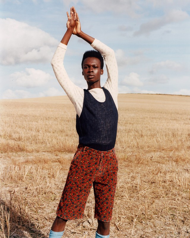 A single dark-skinned model in printed shorts and blue socks standing in a rural field with hands raised in a high fashion pose.