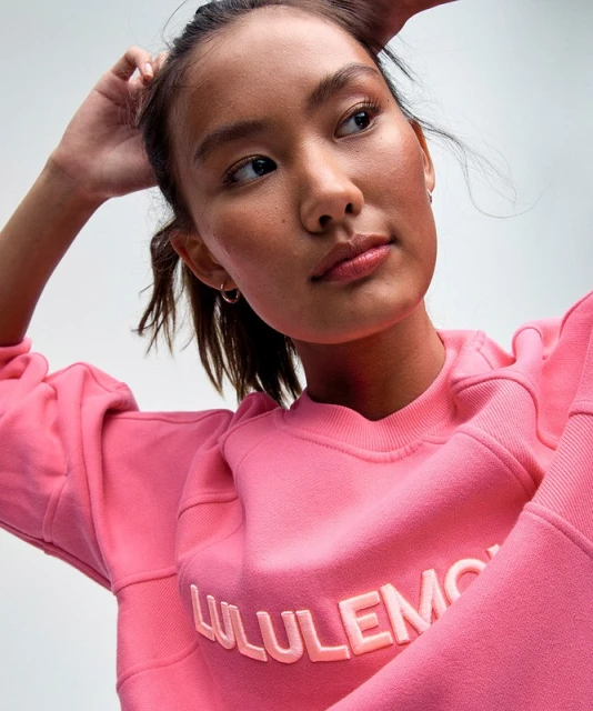 Portrait of an Asian woman in a pink crewneck sweatshirt with 'Lululemon' text, set against a soft white background in a beauty shot.