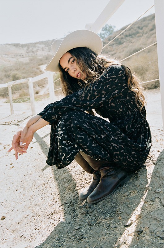 Woman wearing a white cowboy hat and black patterned dress crouching outdoors by a white wooden fence