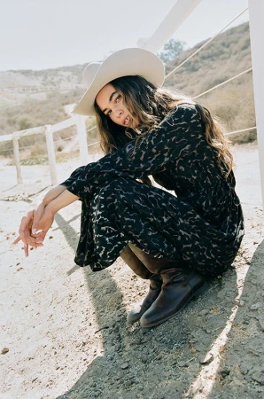 Woman wearing a white cowboy hat and black patterned dress crouching outdoors by a white wooden fence