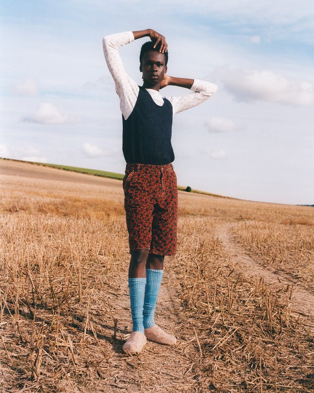 An African male model stands in a harvested field wearing a white shirt, dark vest, patterned shorts, and blue socks, posing with hands up.