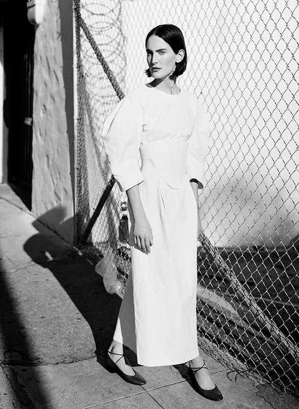Black and white portrait of a woman in a white dress standing beside a chain-link fence in Los Angeles.