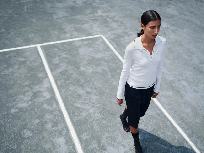 Adult female in casual long-sleeved top and shorts walking on a grey tennis court in elevated outdoor full shot, with light skin and dark hair.
