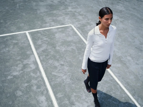 Adult female in casual long-sleeved top and shorts walking on a grey tennis court in elevated outdoor full shot, with light skin and dark hair.
