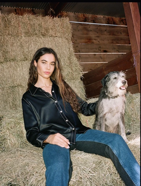 A woman sits on a hay bale with a dog, wearing a black satin shirt, and blue jeans with a wood wall and hay bales behind them.