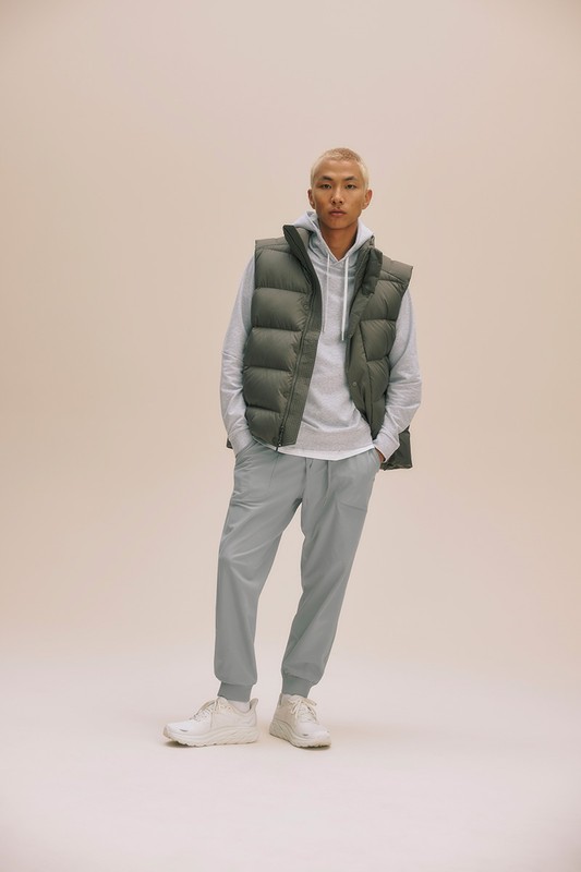 Male model stands in a studio wearing a light blue hooded sweatshirt, green puffer vest, light blue sweatpants, and white sneakers.