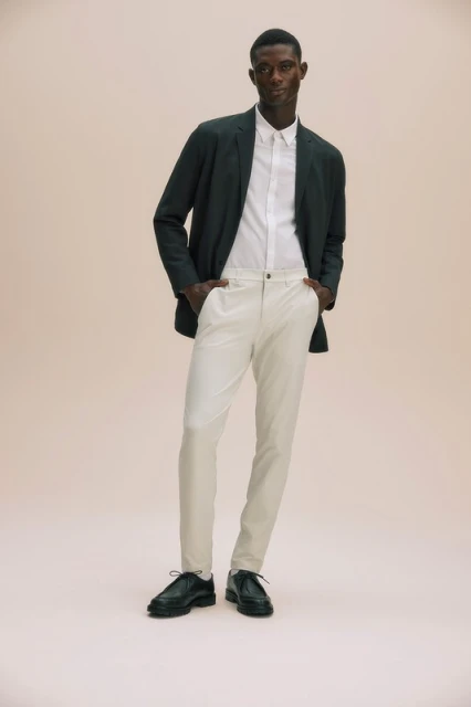 An adult male model poses in a dark green suit jacket, white button-down shirt, and light beige pants in a full studio shot.
