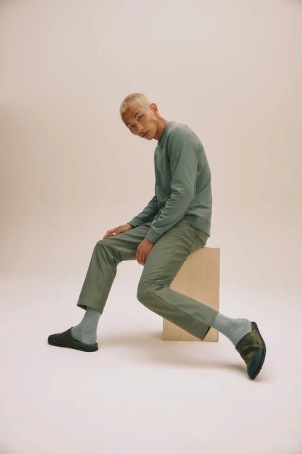 An Asian man with bleached hair sits in a relaxed pose against a neutral backdrop, showcasing a casual sage-green sweatshirt and pants.