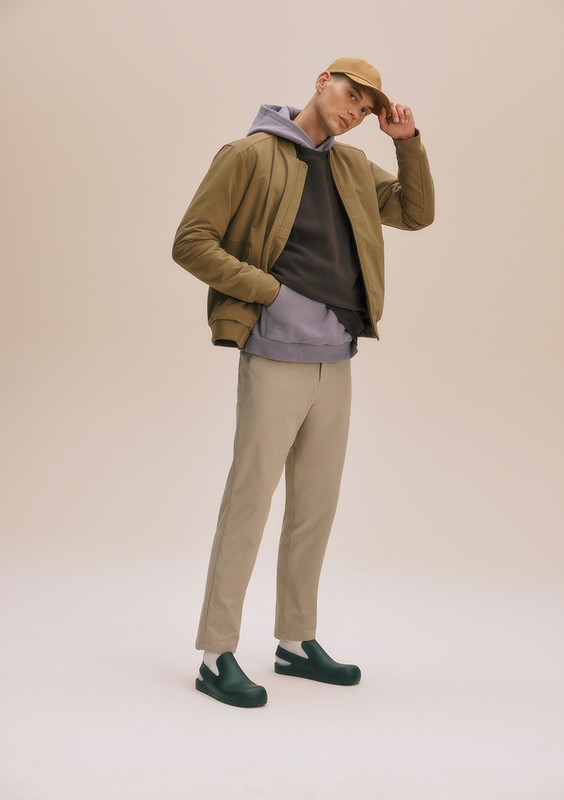 Full-length shot of a man in casual wear posing in front of a neutral backdrop, featuring a jacket, hoodie, trousers, shoes, and a cap.