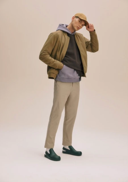Full-length shot of a man in casual wear posing in front of a neutral backdrop, featuring a jacket, hoodie, trousers, shoes, and a cap.
