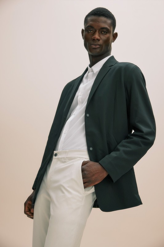 Dark-skinned adult male in a dark blazer, white shirt, and white pants stands indoors with a hand in his pocket, looking at camera.