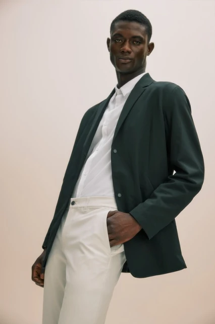 Dark-skinned adult male in a dark blazer, white shirt, and white pants stands indoors with a hand in his pocket, looking at camera.