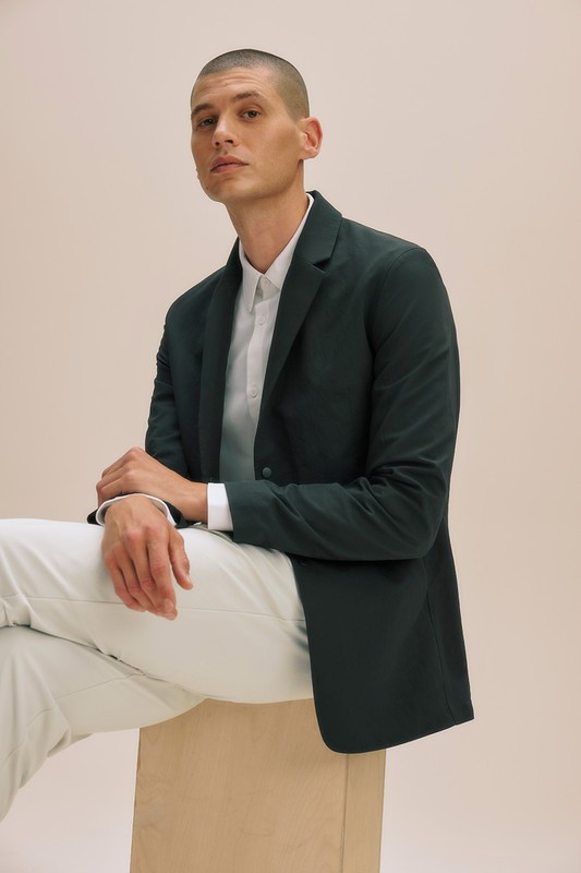 Man in blazer and white shirt sits on box in front of beige backdrop in studio with medium shot. Fashion and lifestyle photography.