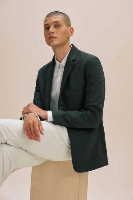 Man in blazer and white shirt sits on box in front of beige backdrop in studio with medium shot. Fashion and lifestyle photography.