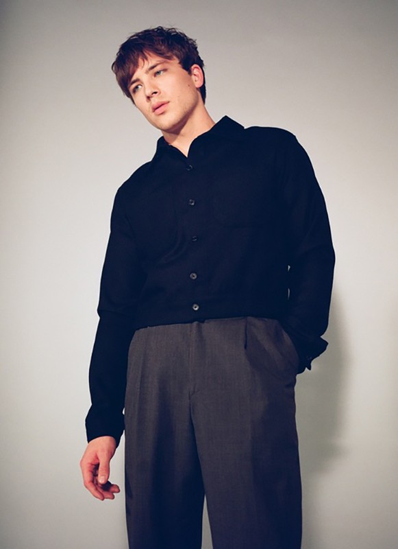 Young adult man in a black button-down shirt and grey slacks poses in a neutral studio setting, gazing off-camera with a slight smile.