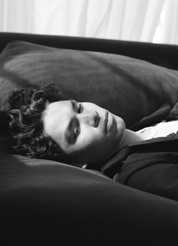 Monochrome portrait of a young adult male with curly dark hair, lying on a dark sofa with white curtains in the background.