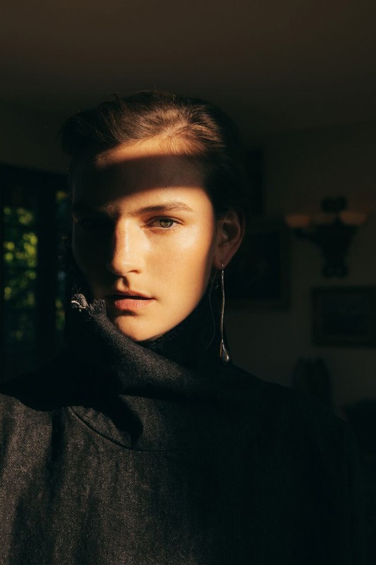 Close-up studio portrait of a fair-skinned woman with earrings wearing a dark turtle neck sweater, half her face lit in golden light.
