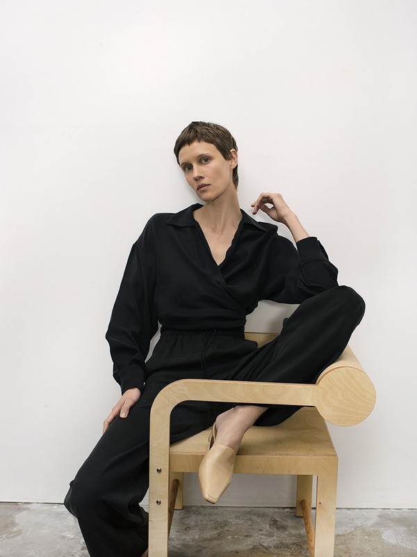 Adult woman in black wrap top and pants seated on a light-wood chair in front of a white wall in a minimalist studio setting.