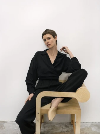 Adult woman in black wrap top and pants seated on a light-wood chair in front of a white wall in a minimalist studio setting.