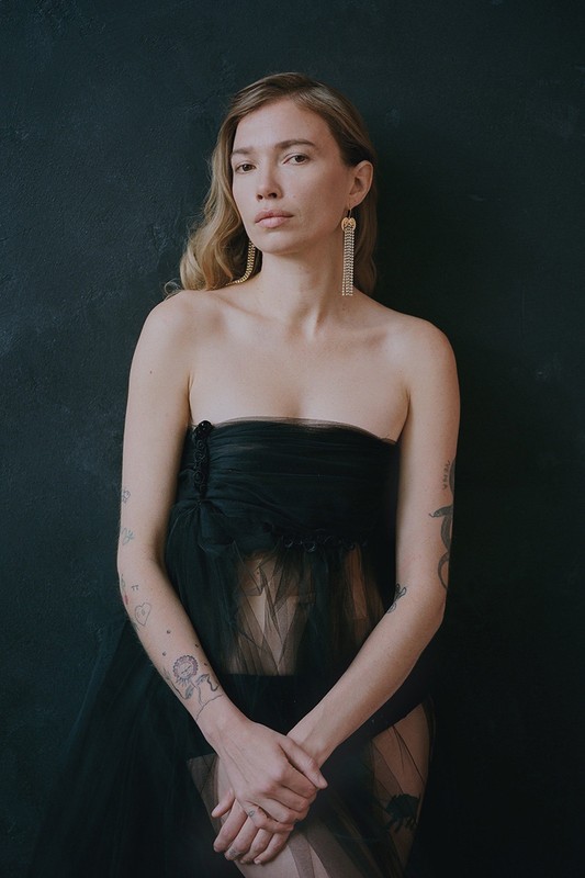 Portrait of an adult female with blonde hair, wearing a black strapless dress and long earrings, posing against a dark background.