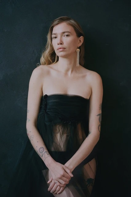 Portrait of an adult female with blonde hair, wearing a black strapless dress and long earrings, posing against a dark background.