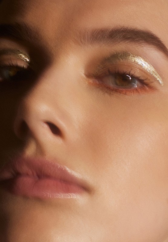 Close-up portrait of a woman with gold eyeshadow. Her face is partially visible, soft focus on skin texture and eye detail.